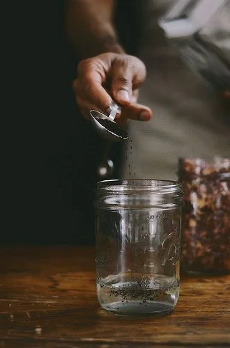 A hand holding a spoon above a glass mason jar, pouring black pepper into the jar on a wooden surface.