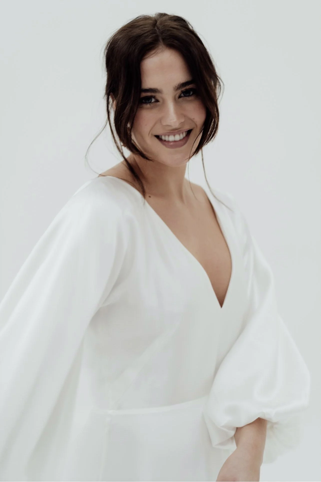 Smiling woman with dark brown hair styled in loose waves, wearing a white dress with a V-neck and puffed sleeves, standing against a plain white background.