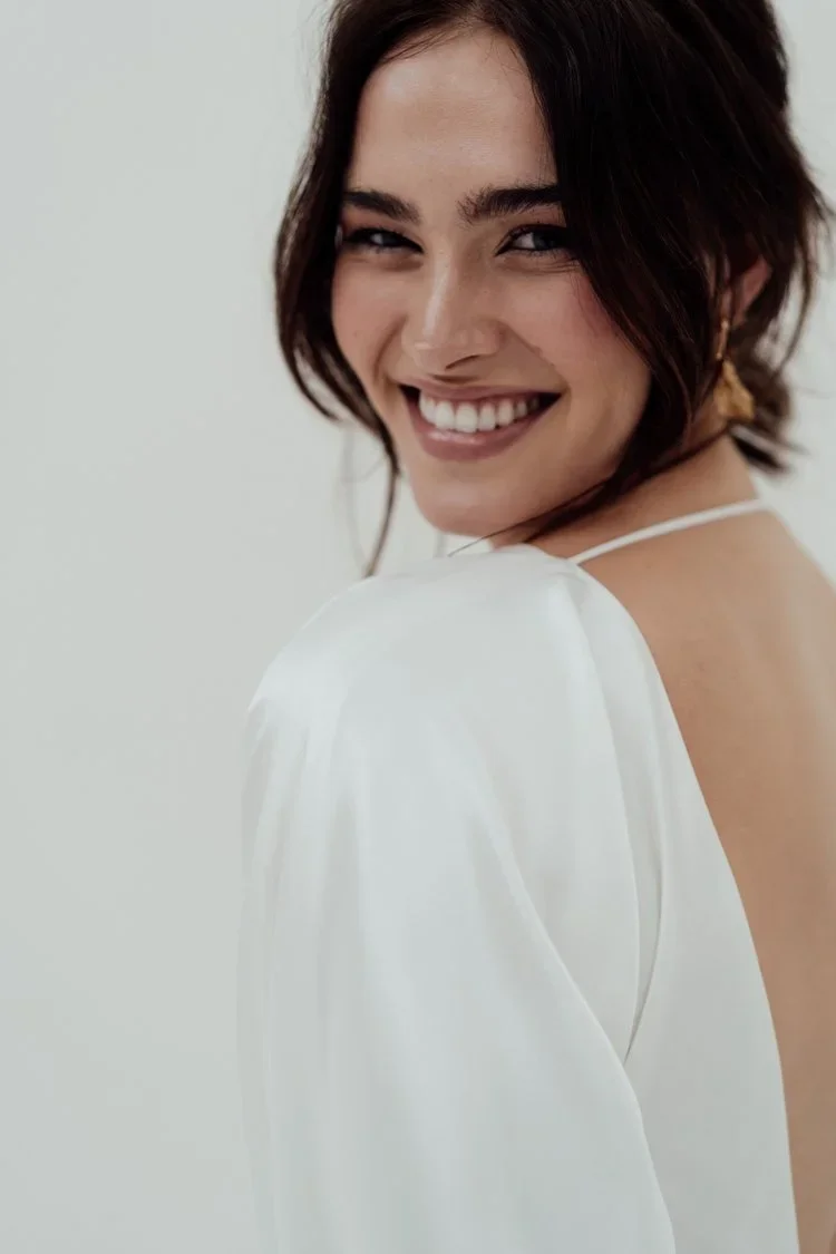 A radiant bride wearing natural ethical bridal makeup, glowing skin with a soft dewy finish, subtle champagne shimmer on the eyelids, softly defined brows, delicate rose-toned blush, and a muted petal-pink lip. Her complexion looks fresh breathable.
