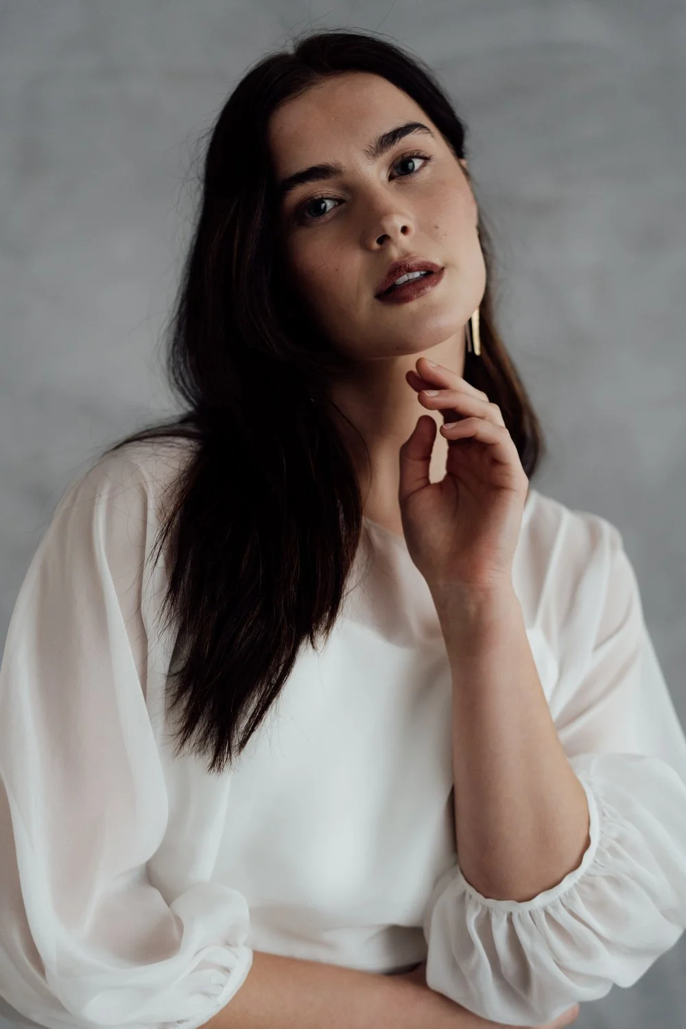 A young woman with long dark hair, wearing a white blouse and gold hoop earrings, looking at the camera with a gentle expression, her hand resting near her chin, against a neutral background.