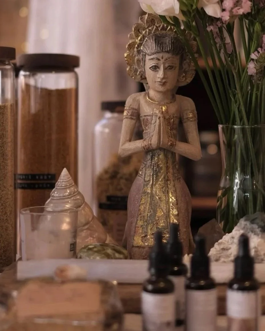 A decorative wooden figurine of a woman in traditional attire with hands in a prayer position, surrounded by glass bottles, jars, flowers, and various objects on a table.