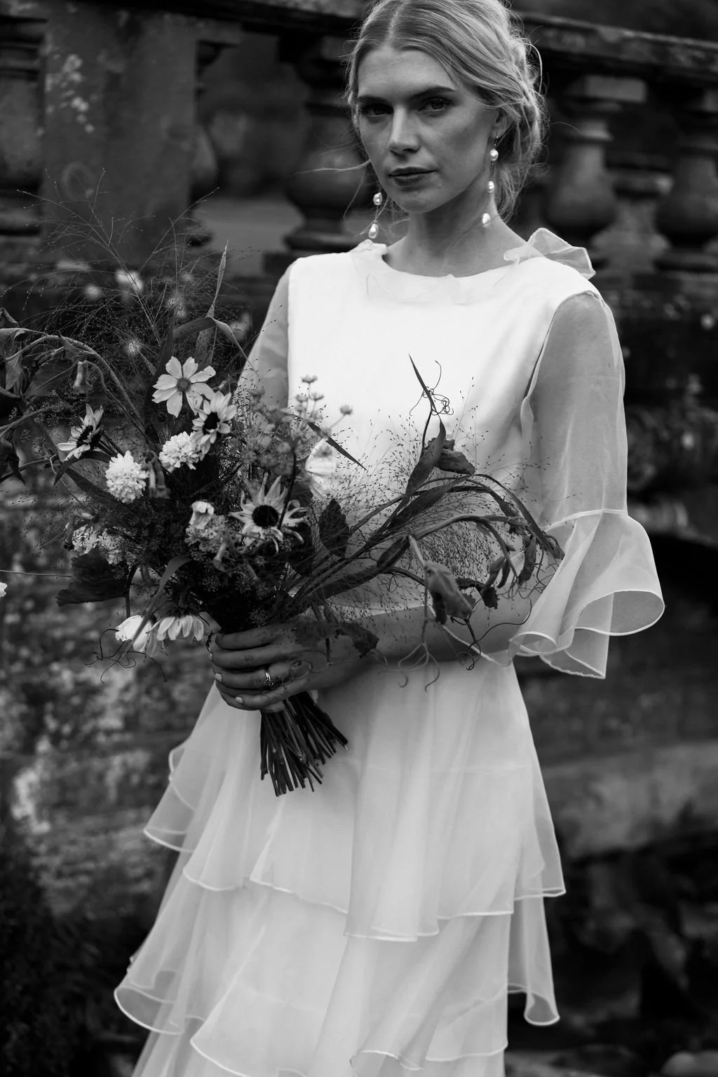 A woman in a white dress holding a bouquet of flowers, standing outdoors near a stone structure.