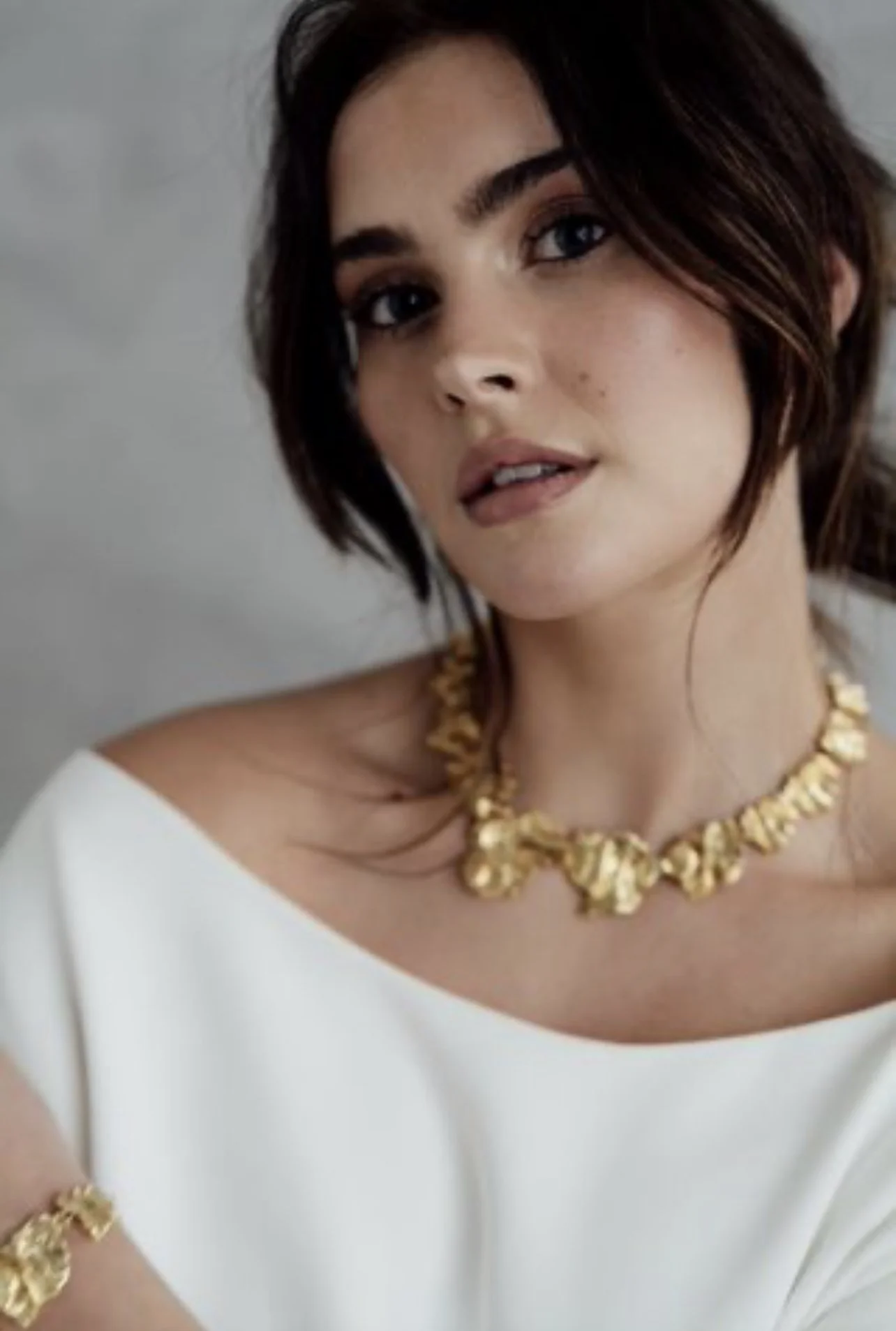 A woman with short dark hair and fair skin wearing a white top, gold jewelry including a necklace and bracelet, and looking at the camera with a slight smile.