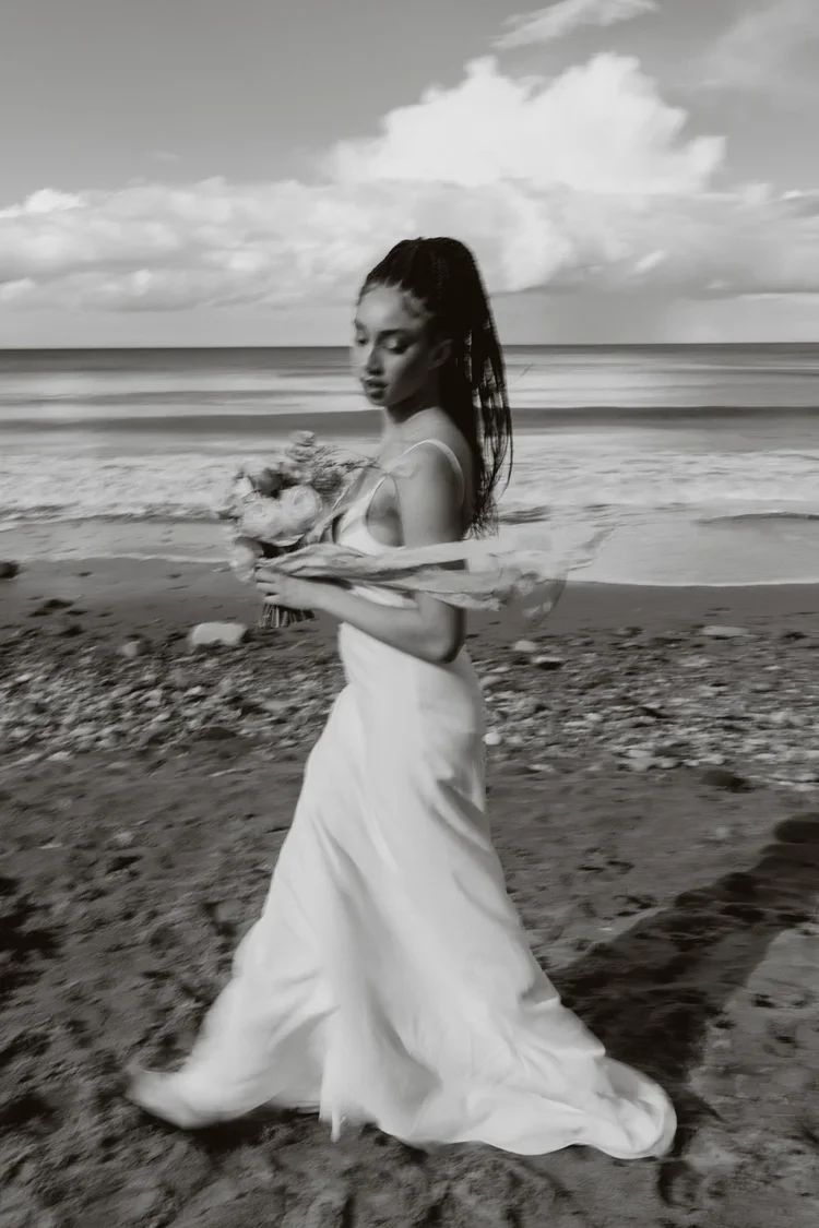 A woman wearing a long dress holding a bouquet of flowers walking along a beach with ocean waves and clouds in the background.