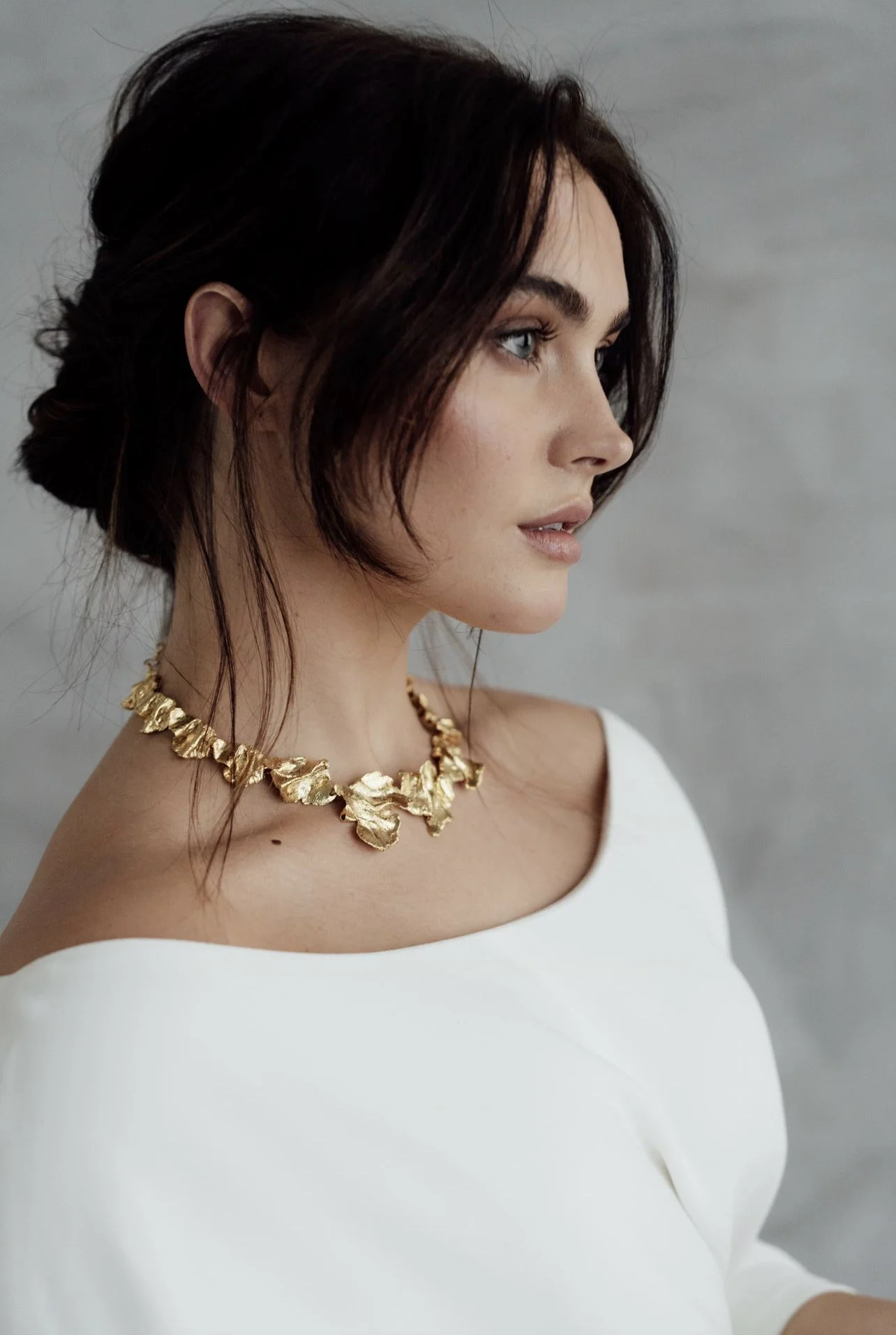 A woman with dark brown hair styled in a loose updo, wearing a white off-shoulder top and a gold leaf necklace, looking to the side against a gray background.