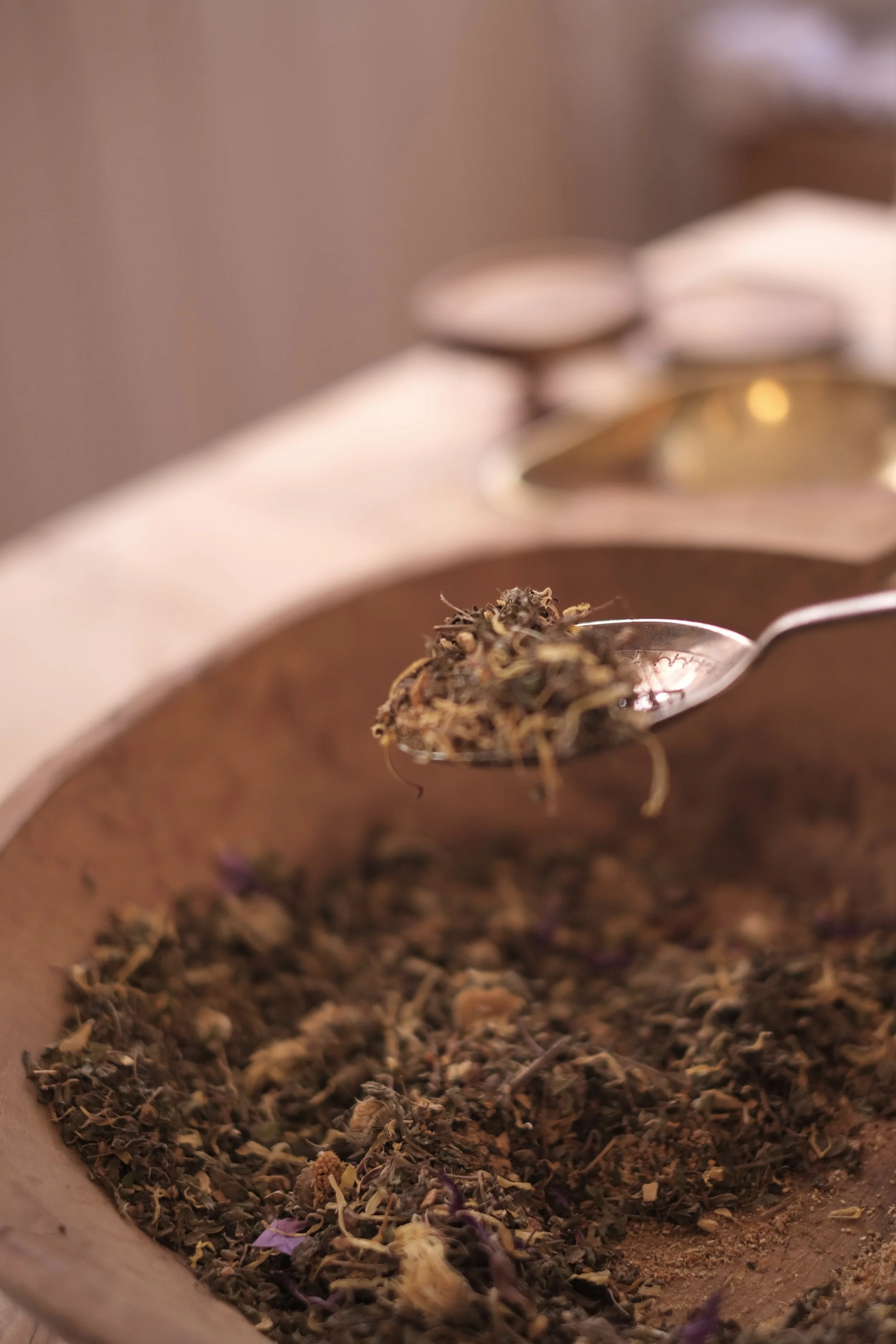 Close-up of a spoonful of dried herbal tea being held above a wooden bowl filled with more dried herbs, with some gold-colored containers in the blurry background.