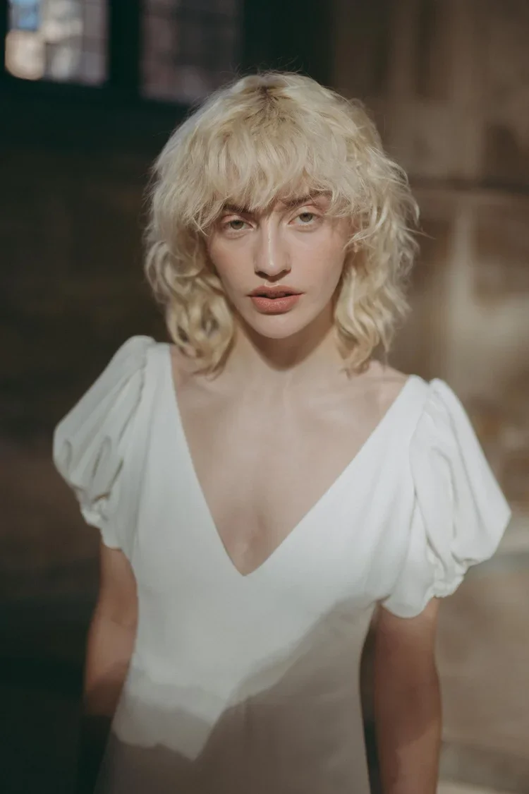 A woman with curly blonde hair and light skin wearing a white top with puffed sleeves, looking directly at the camera in an indoor setting.
