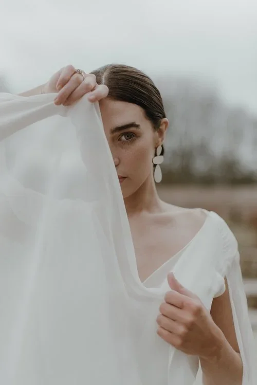 A woman with dark hair and earrings holding a white fabric, partially covering her face outdoors.