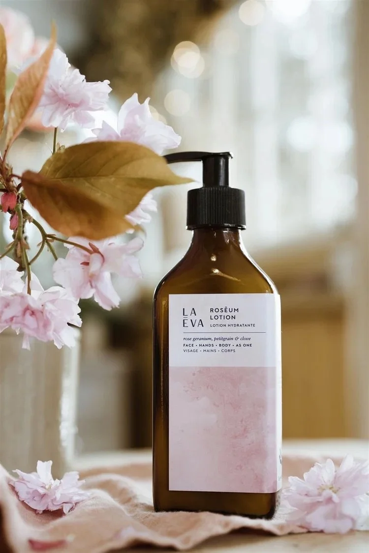 A brown bottle of La Leva roseum lotion with a black pump, placed on a soft pink cloth next to pink flowers and leaves, in a softly lit room.
