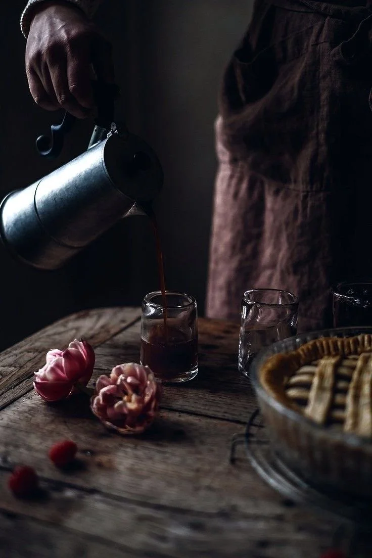 Hand pouring coffee from a metal pot into a glass on a rustic wooden table, with flowers and a bowl of cookies nearby.