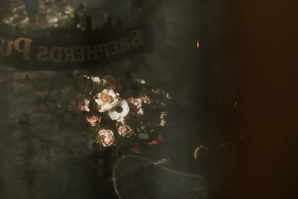 A bouquet of artificial or real flowers, including roses and anemones, seen through a foggy or reflective surface with a blurred background that appears to include a banner with partially visible text.