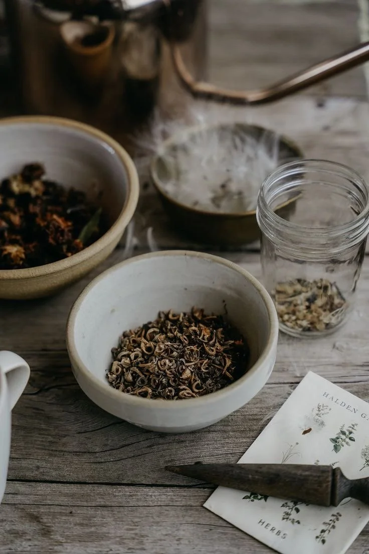 Bowls of dried herbs and flowers, a jar with herbs, a small bowl with white foam or powder, a paper with botanical illustrations, and a rustic wooden surface.