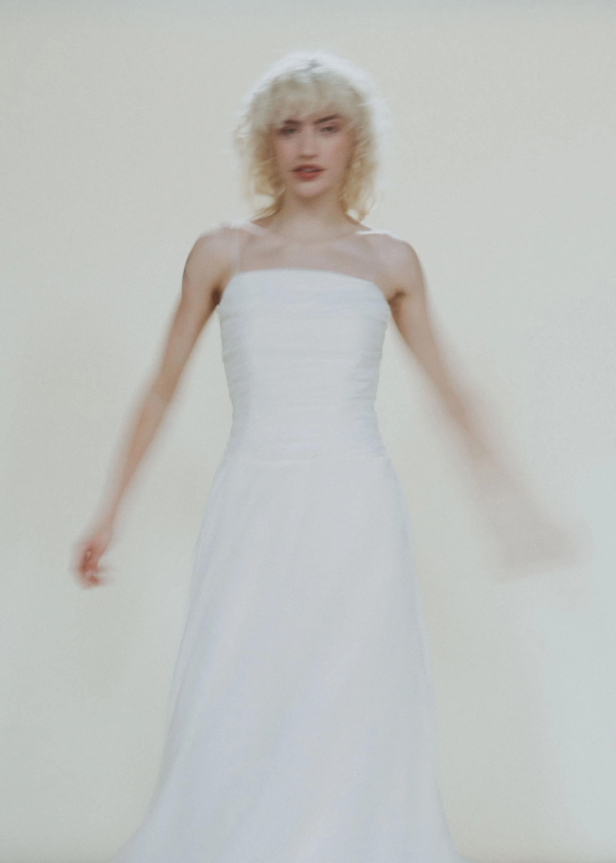 A woman with short, curly blonde hair wearing a white strapless dress stands against a plain background.