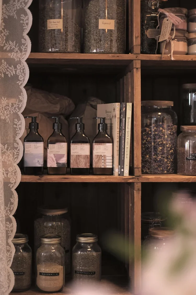 Wooden shelves with jars containing herbs and spices, books about botanicals, and labeled bottles of hair care products.