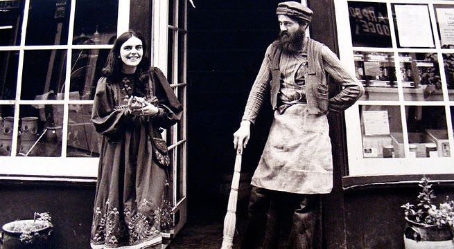 A young woman and an older man dressed in vintage clothing stand outside a shop or café with large windows. The woman is smiling, while the man has a neutral expression, holding a cane or walking stick. The background shows reflections in the windows