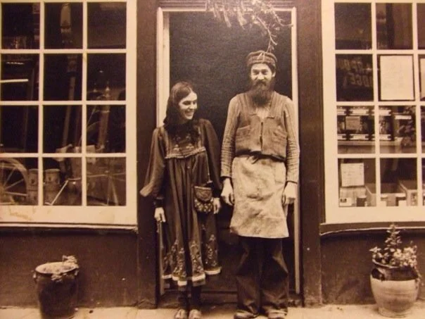 A woman and a man dressed in old-fashioned clothing standing in a doorway outside of a building with large windows. The woman is wearing a long dress, and the man is wearing a shirt, apron, and turban.