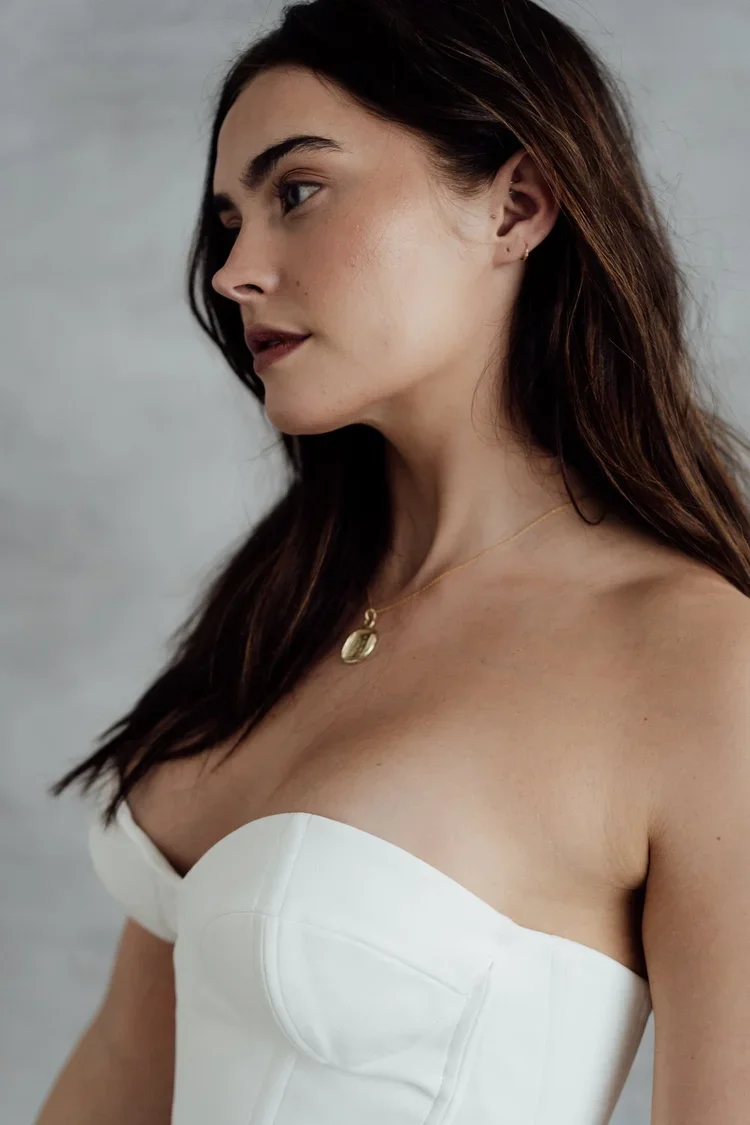 A woman with long brown hair wearing a strapless white top and a gold pendant necklace, looking to her left.