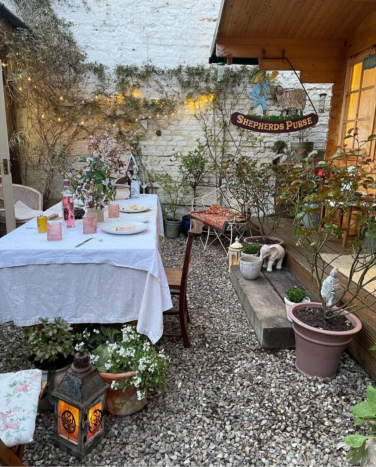 An outdoor patio decorated for a gathering with a long white table, pink glasses, bottles, plates, and floral centerpieces. Potted plants and small decorative statues decorate the area, with a brick wall and string lights overhead. A sign reads 'Shep