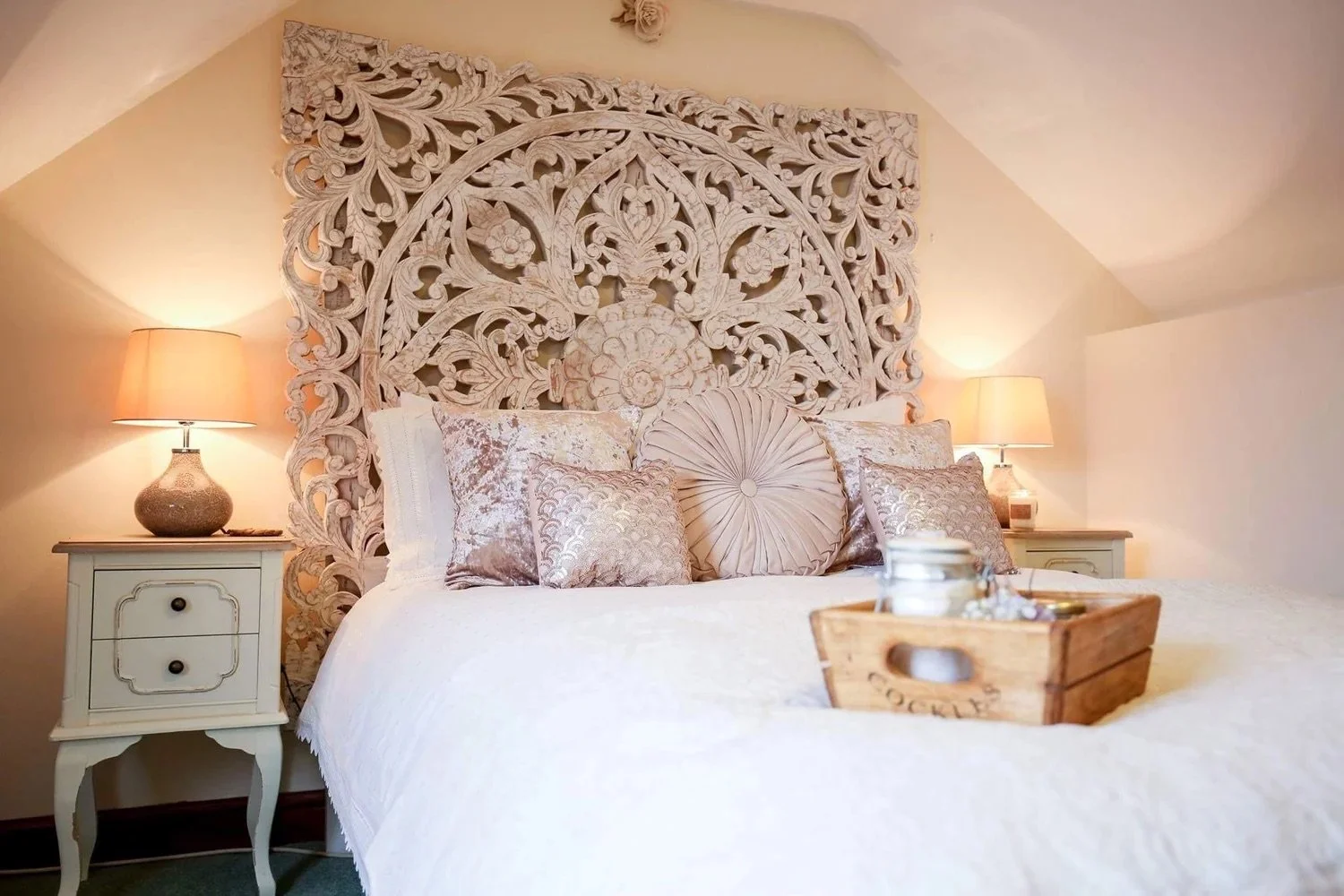 A cozy bedroom with a large ornate wooden headboard, nightstands with lamps, and plush pillows on a white bedspread. A wooden tray with candles and decorative items is on the bed.