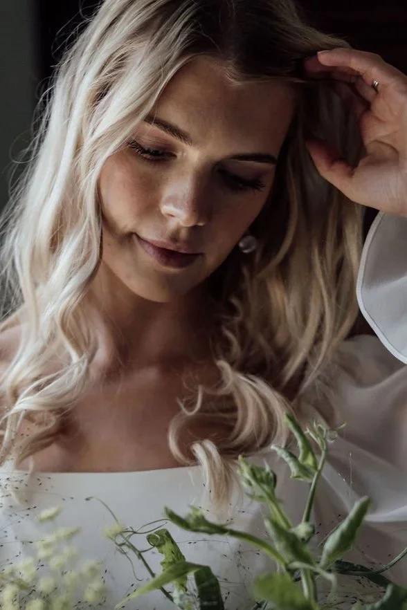 A woman with long blonde hair, eyes closed, gently touching her hair, with a soft smile. She is wearing a white top and holding a bouquet of green plants.