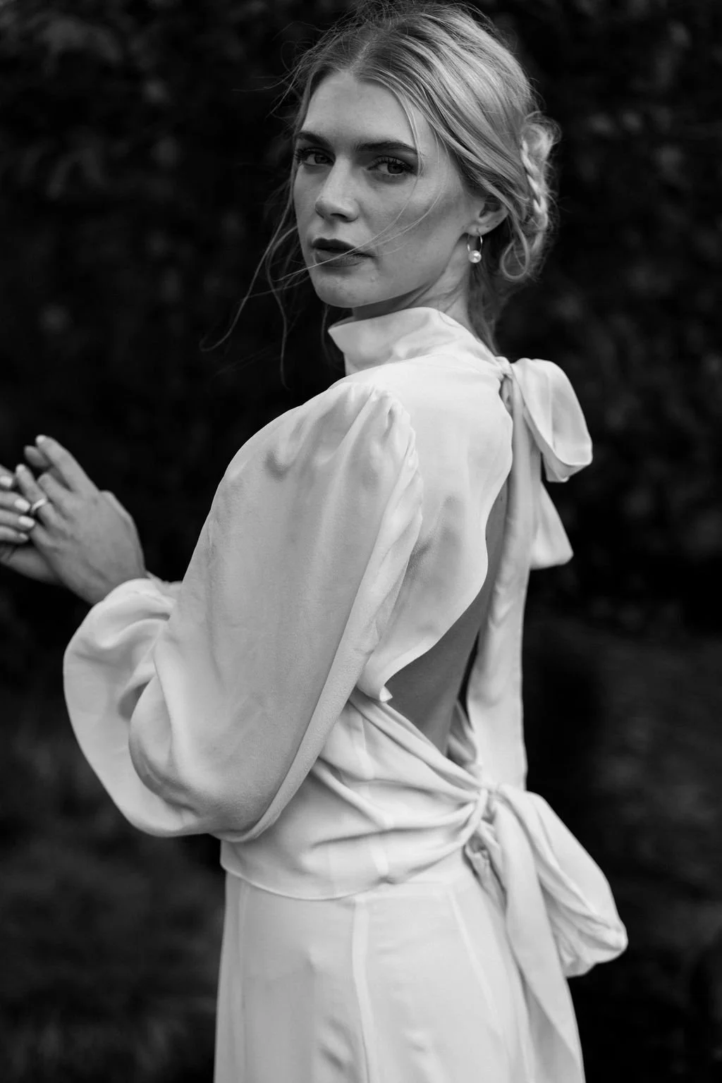 A woman with light-colored hair styled in an updo, wearing elegant earrings and a smooth, silky blouse with puffed sleeves and a bow at the back, standing outdoors with a blurred dark background.