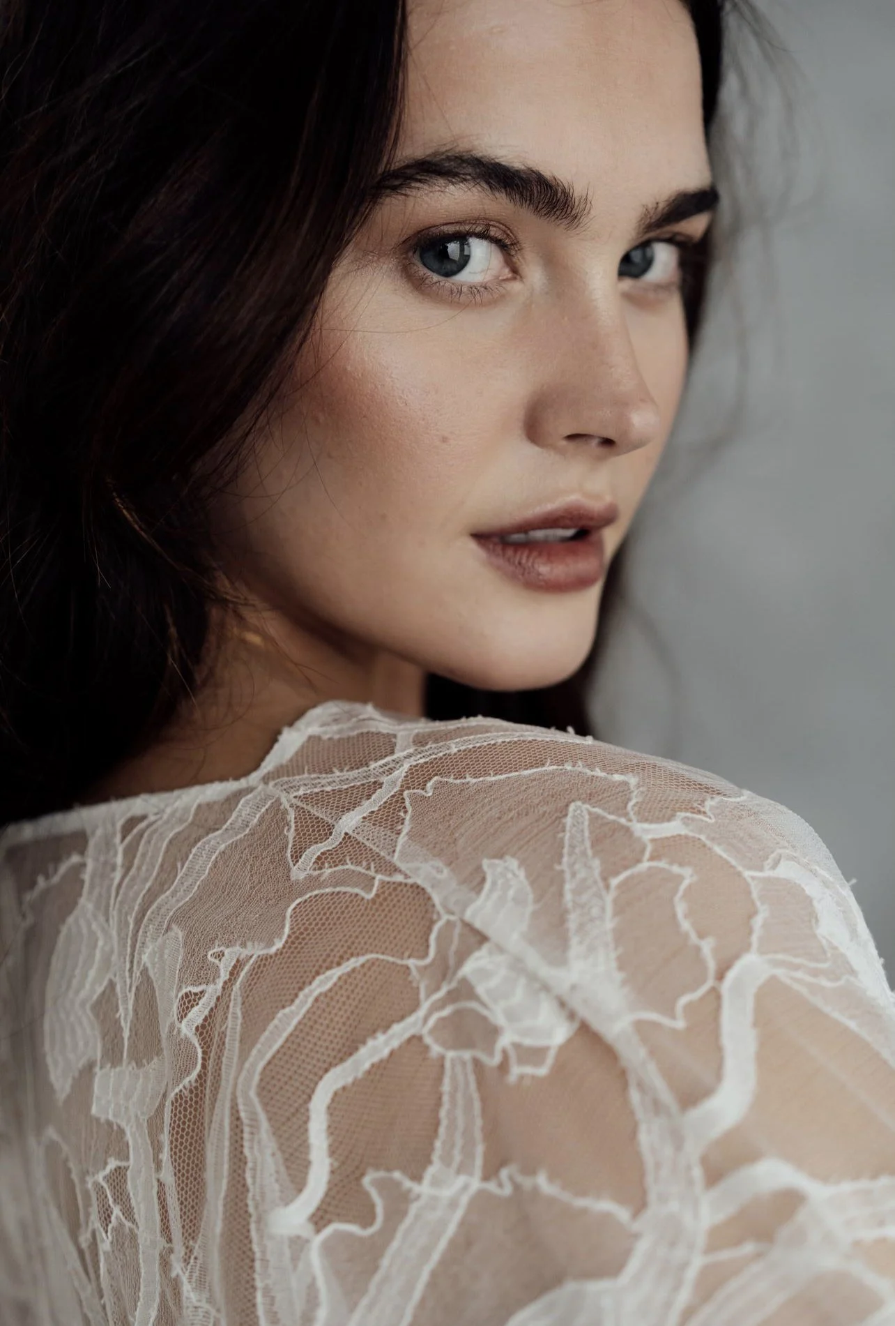 Close-up of a woman with dark brown hair, blue eyes, and fair skin looking over her shoulder, wearing a sheer, embroidered white lace top.