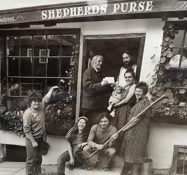 A group of people, including two women, three children, a man, and a woman holding a baby, outside a shop named Shepherds Purse, with one woman holding a watering can and a woman holding a broom.