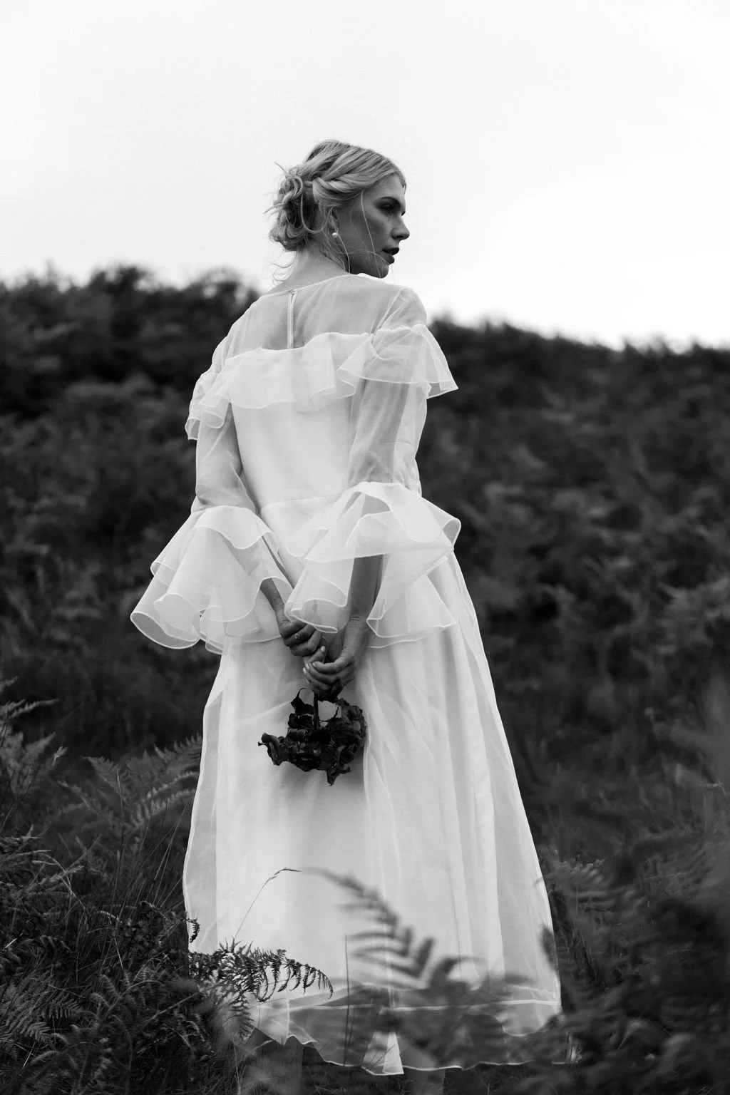 A woman in a flowing, ruffled dress holding a small bouquet of flowers stands outdoors in a natural, grassy landscape, captured in black and white.