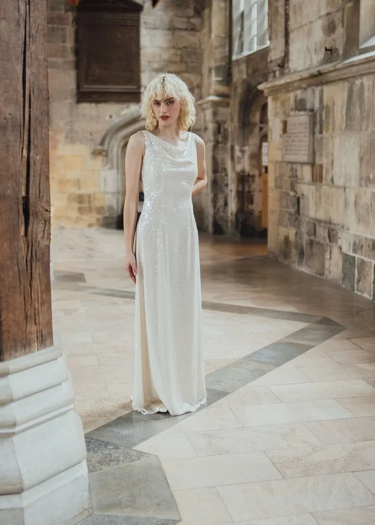 A woman with blonde curly hair in a white, sparkly gown standing inside a historic stone building with arched windows and wooden beams.