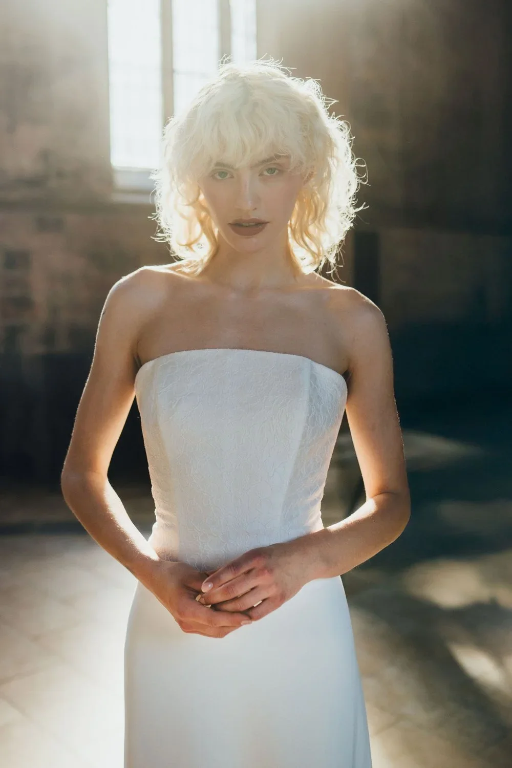 A woman with curly blonde hair in a white strapless dress standing in a sunlit industrial-style room with brick walls and large windows.