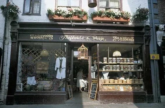 Front of a store called Shepherd's Deli with window display of clothes, and shelves with jars, with flowers and windows above.