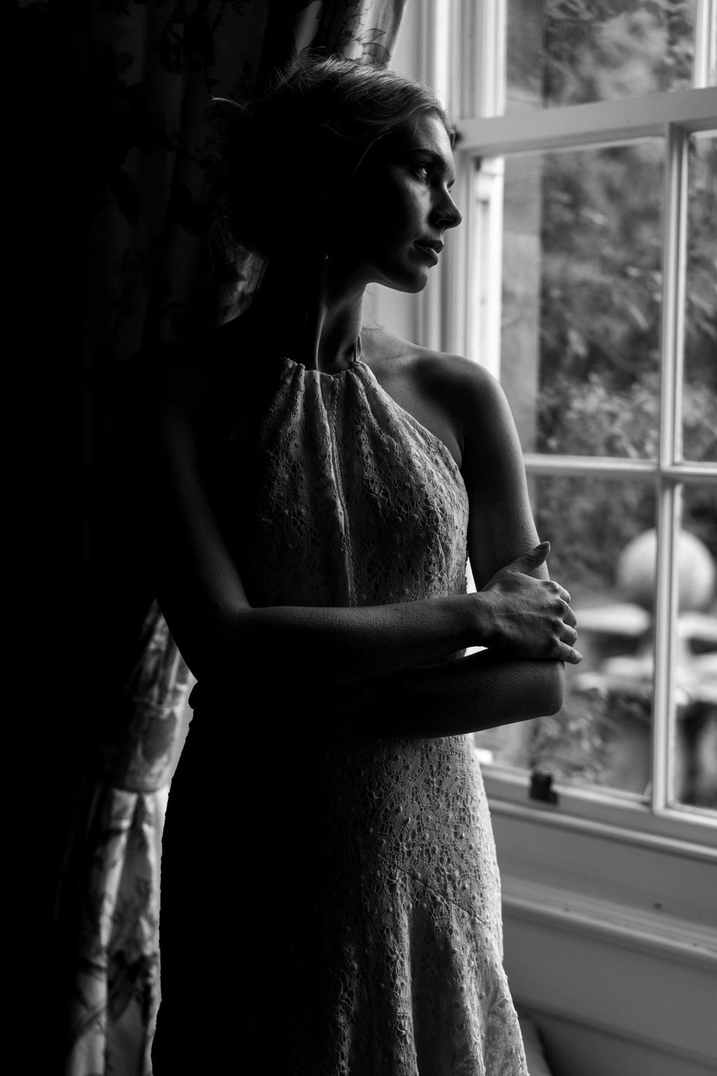 A black and white photo of a woman standing by a window, looking outside. She has a contemplative expression, her arms crossed, wearing a sleeveless dress with lace details, and the light highlights her profile.