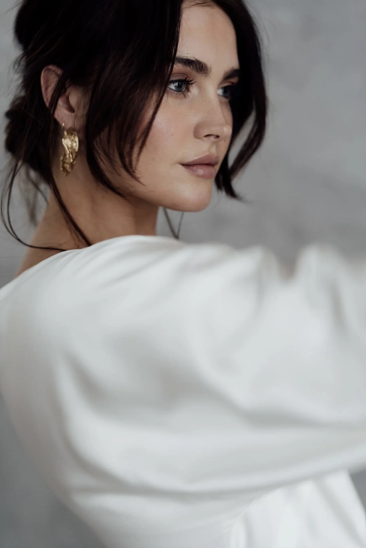 Close-up portrait of a woman with short dark hair, blue eyes, and gold earrings, wearing a white top, against a plain background.