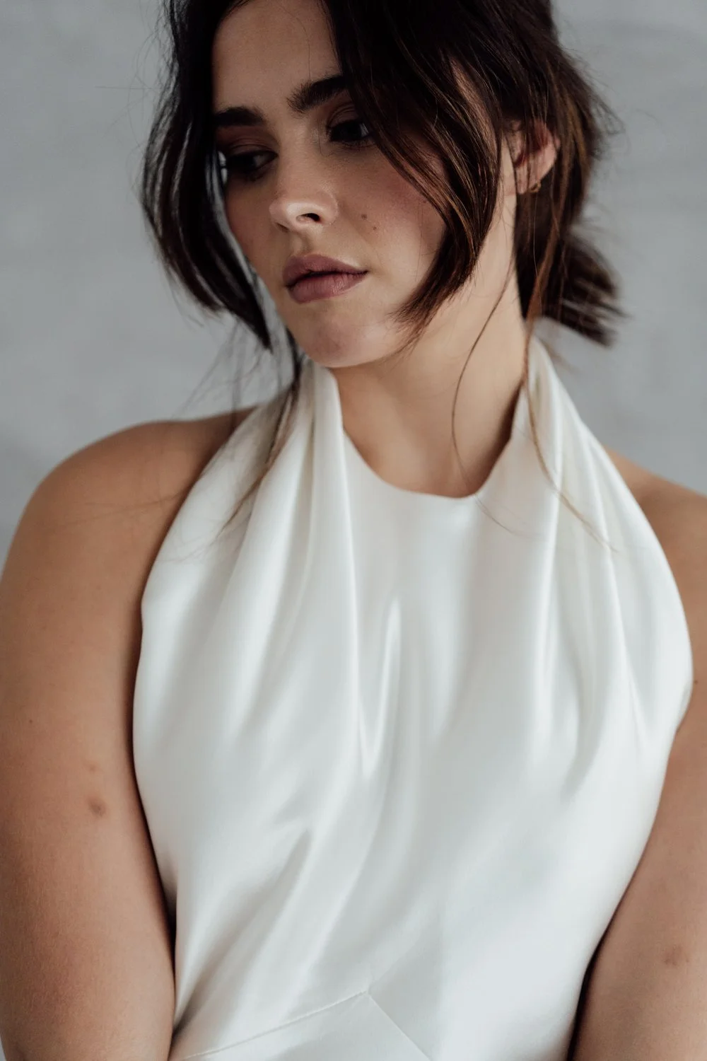 A woman with short, dark brown hair and light skin, wearing a sleeveless white satin top, looking down with a neutral expression.