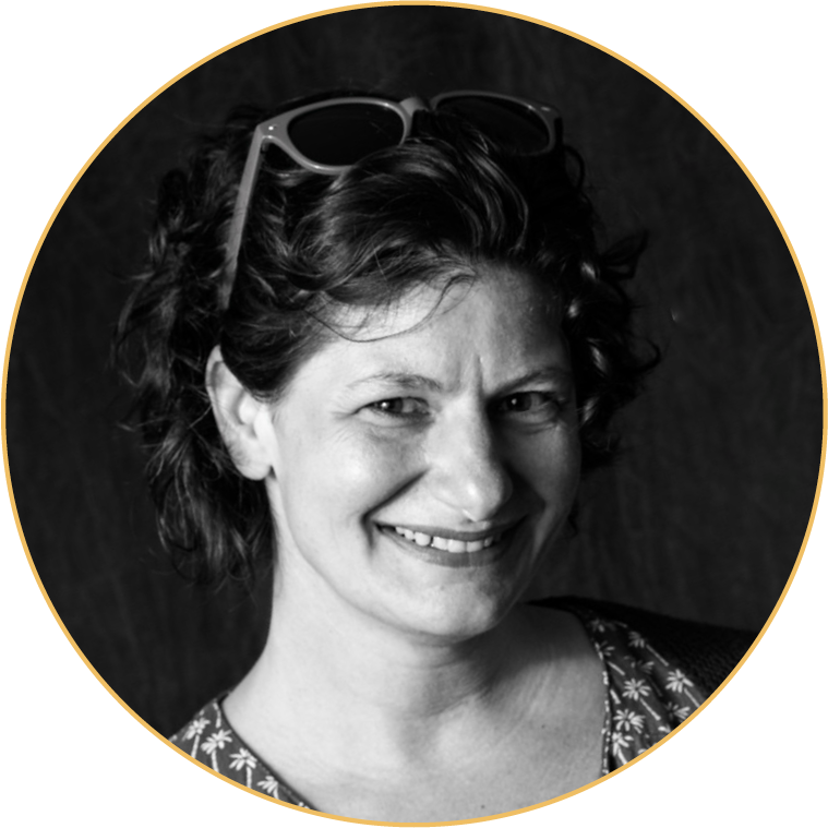 A black-and-white portrait of a woman smiling, with short curly hair, eyeglasses resting on her head, wearing a patterned top, against a dark background.