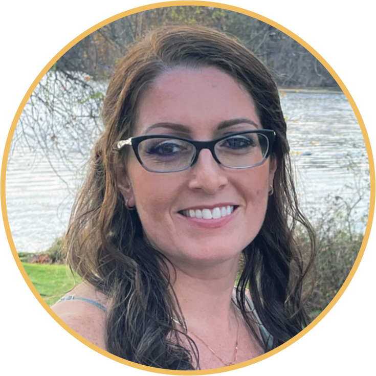 A woman with glasses smiling outdoors near a body of water, with trees in the background.