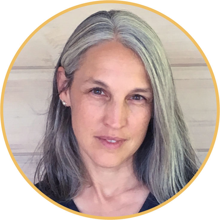 A woman with long gray hair and light skin, smiling slightly, in front of a light-colored wooden background.