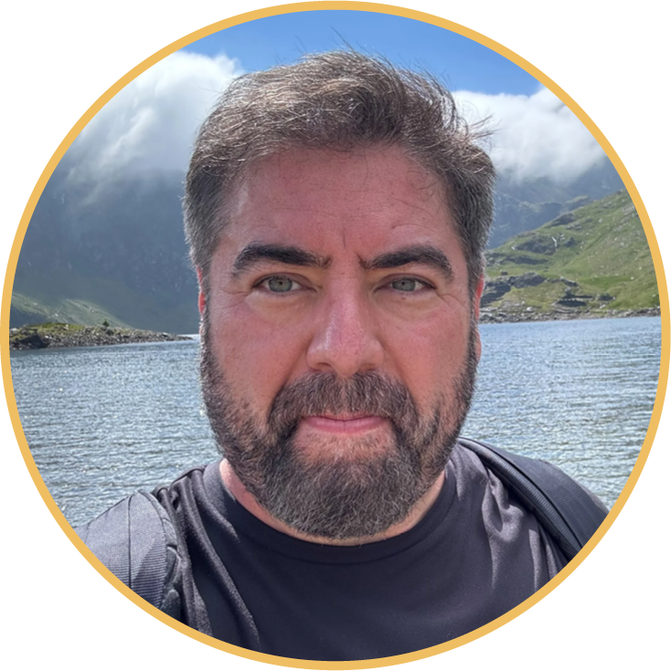 A man with a beard and mustache taking a selfie by a lake with mountains and cloudy sky in the background.