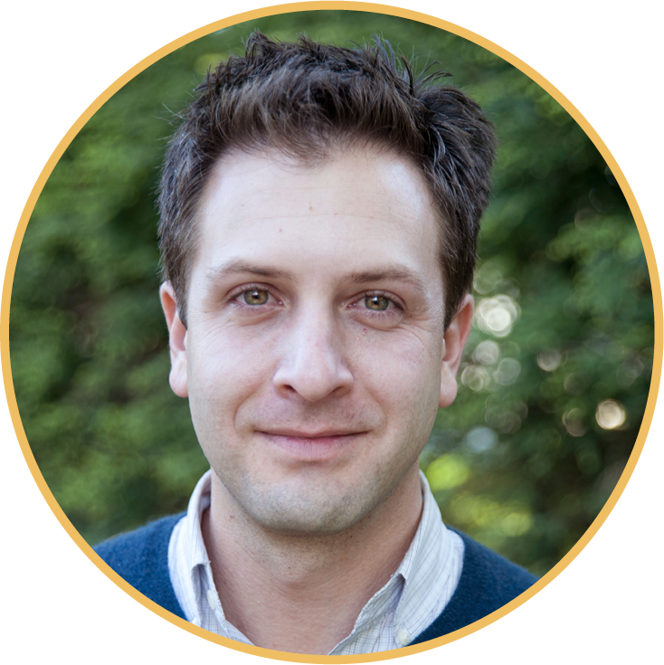 Headshot of a young man with short, dark hair, smiling softly, outdoors with a blurred green background.
