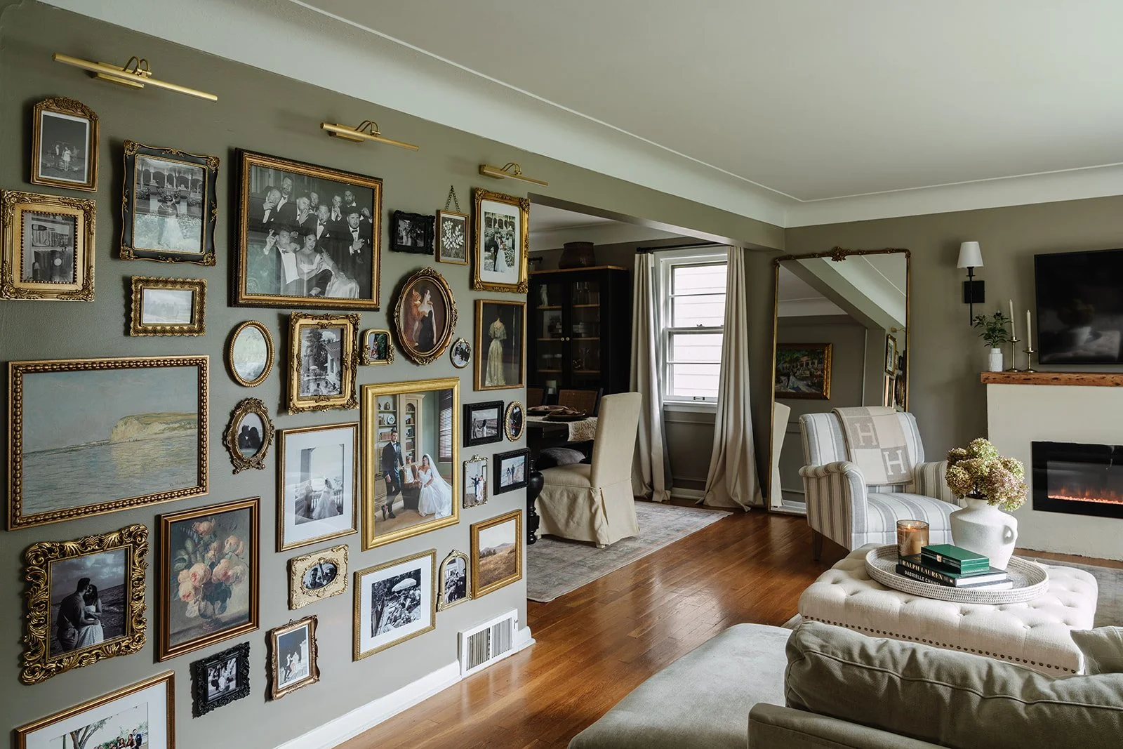 Living room with a wall decorated with many picture frames, a fireplace, white armchair with striped fabric, a tufted ottoman with flowers and books, and a gallery wall with a large mirror and window in the background.