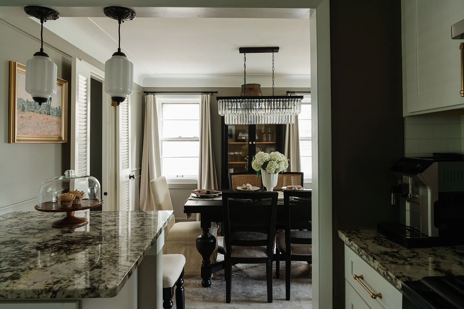A cozy dining area seen from the kitchen doorway with a dark wood table, white flowers in a vase, and large windows with white curtains.