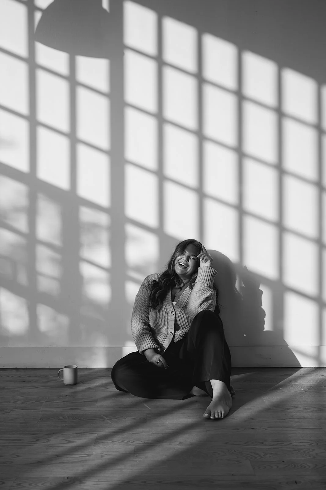 A woman sitting on a wooden floor, smiling and laughing, with sunlight and shadows from a window grid casting patterns on the wall behind her.