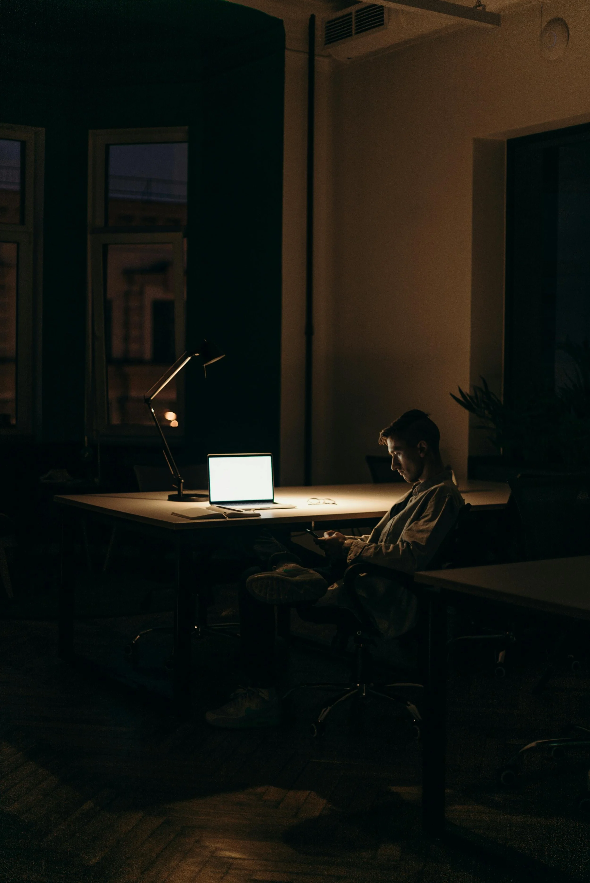 A young man sitting at a desk in an office at night, working on his phone with a laptop and desk lamp on the desk. The office is dimly lit, with a window showing a dark outside.