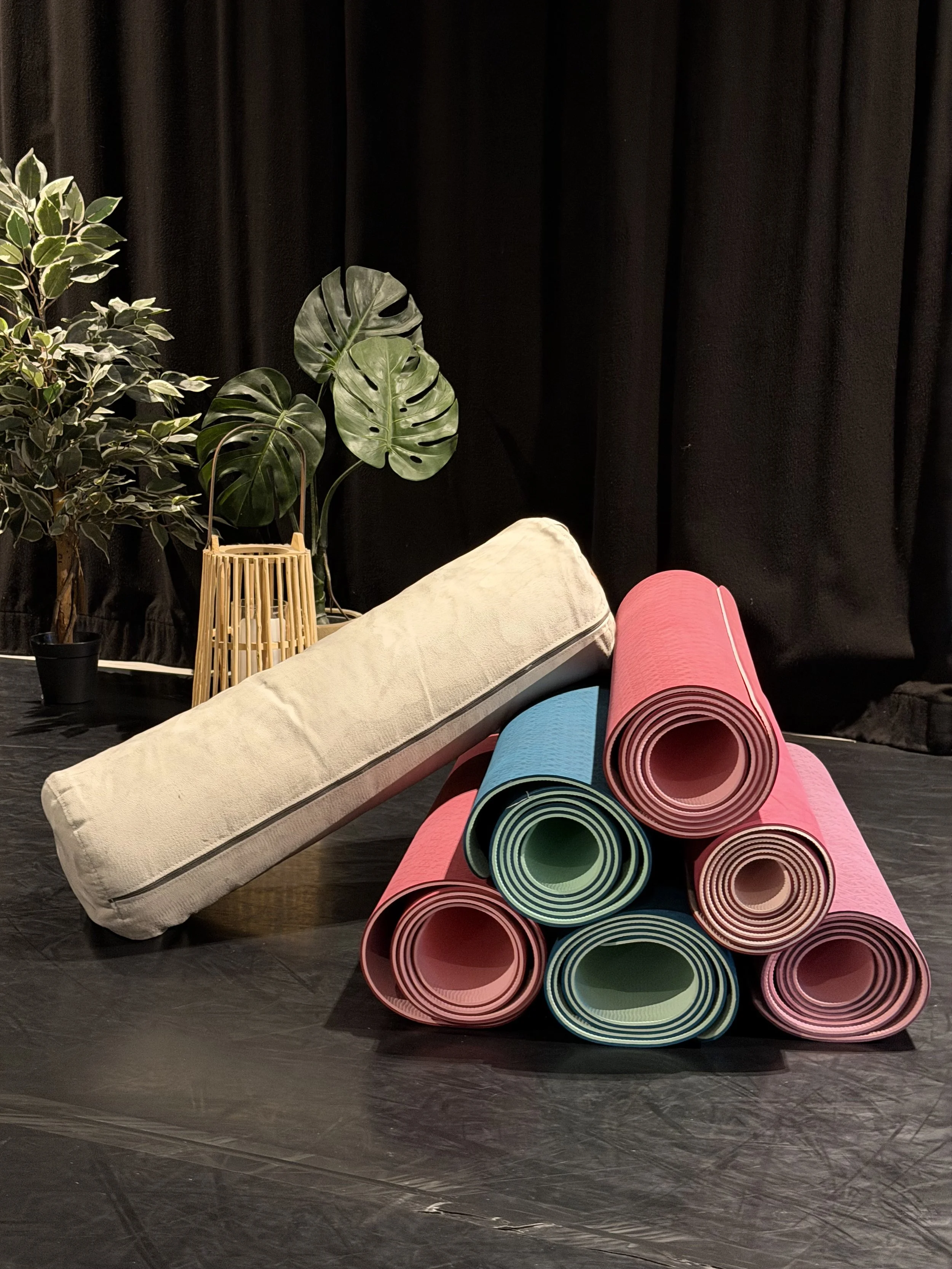Stacked colorful yoga mats on black floor in front of a black curtain, with potted plants and a woven decorative object in background.