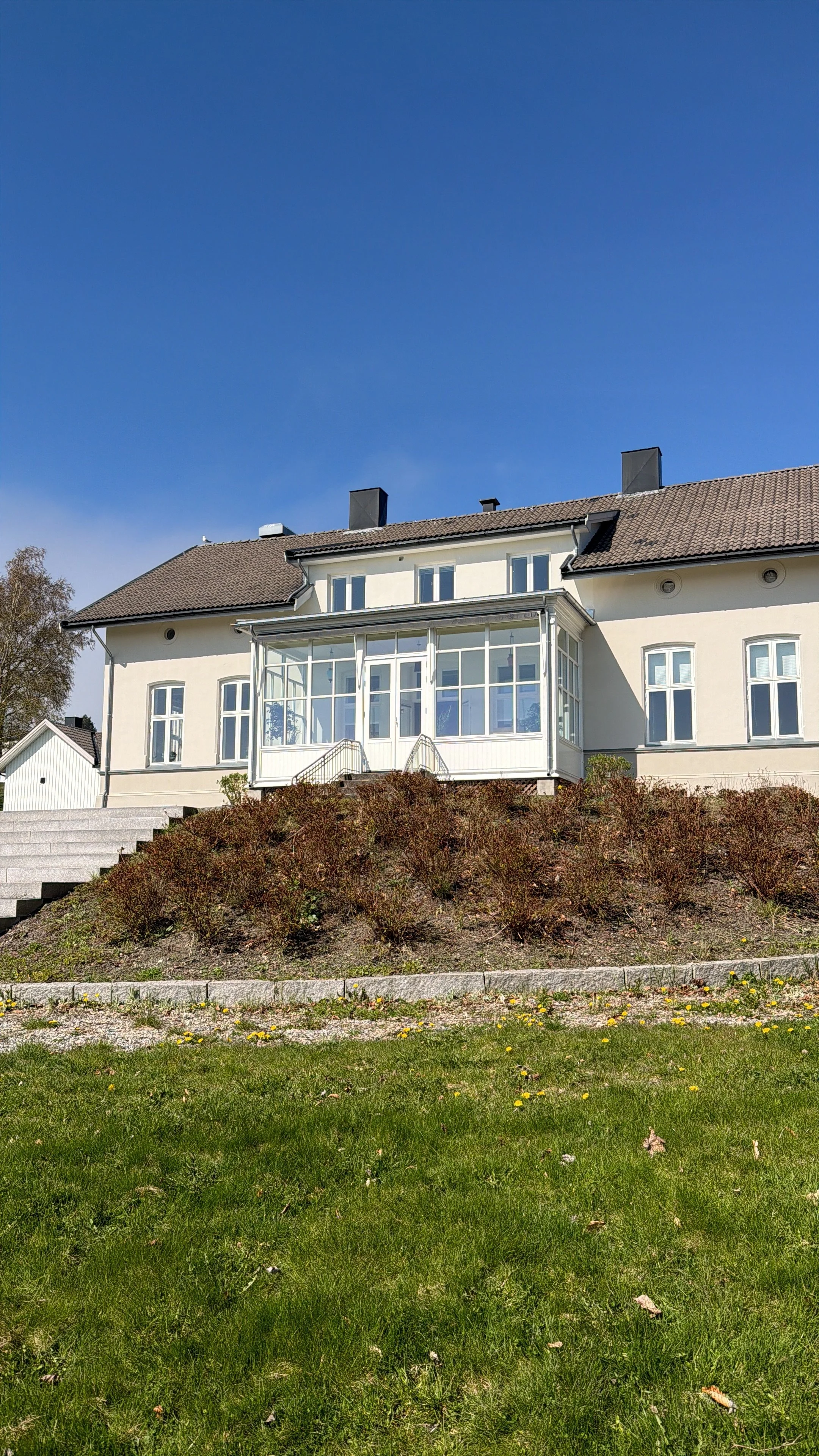 Hus med hvit utvendig veranda og trapp, omgitt av hage med blomster og busker, under en klar blå himmel.