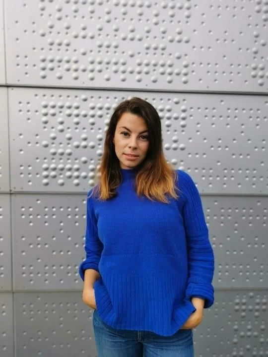 Young woman with shoulder-length brown hair wearing a blue sweater and jeans, standing against a gray textured wall.