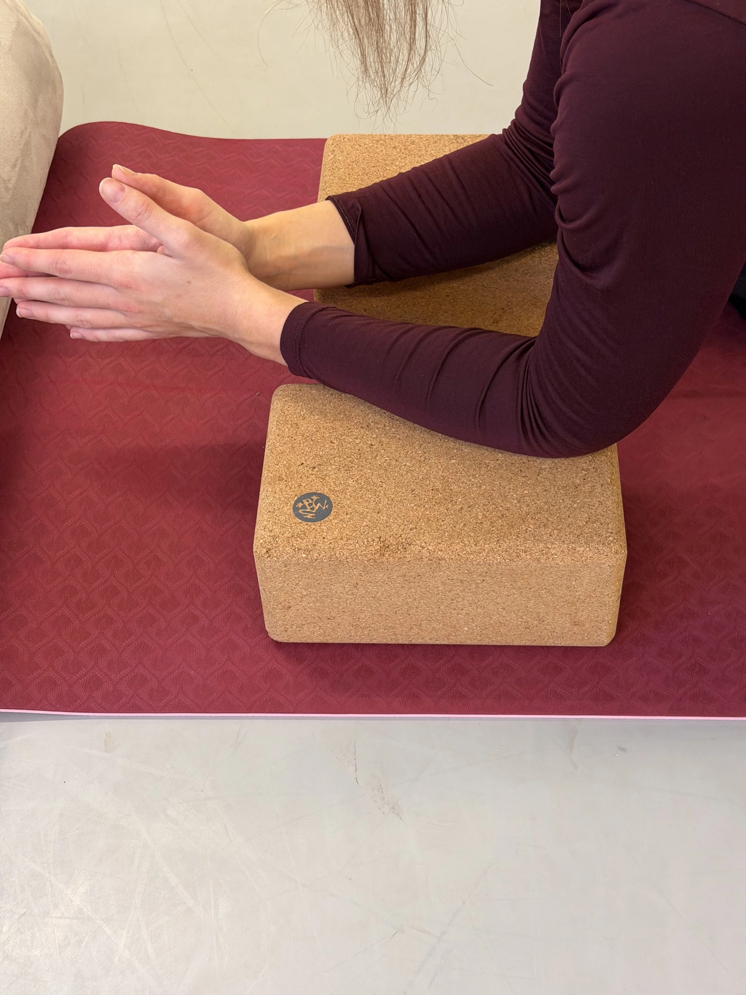 Person practicing yoga or stretching with hands pressed together on a cork yoga block, on a purple mat, with a maroon long sleeve shirt.