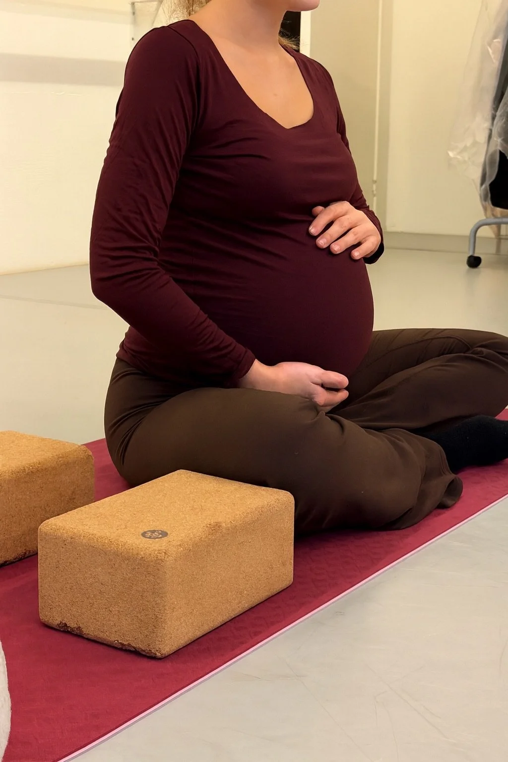 Kvinne i rolig yoga-stilling, sittende på knærne på yogamatte med tregårder rundt seg.