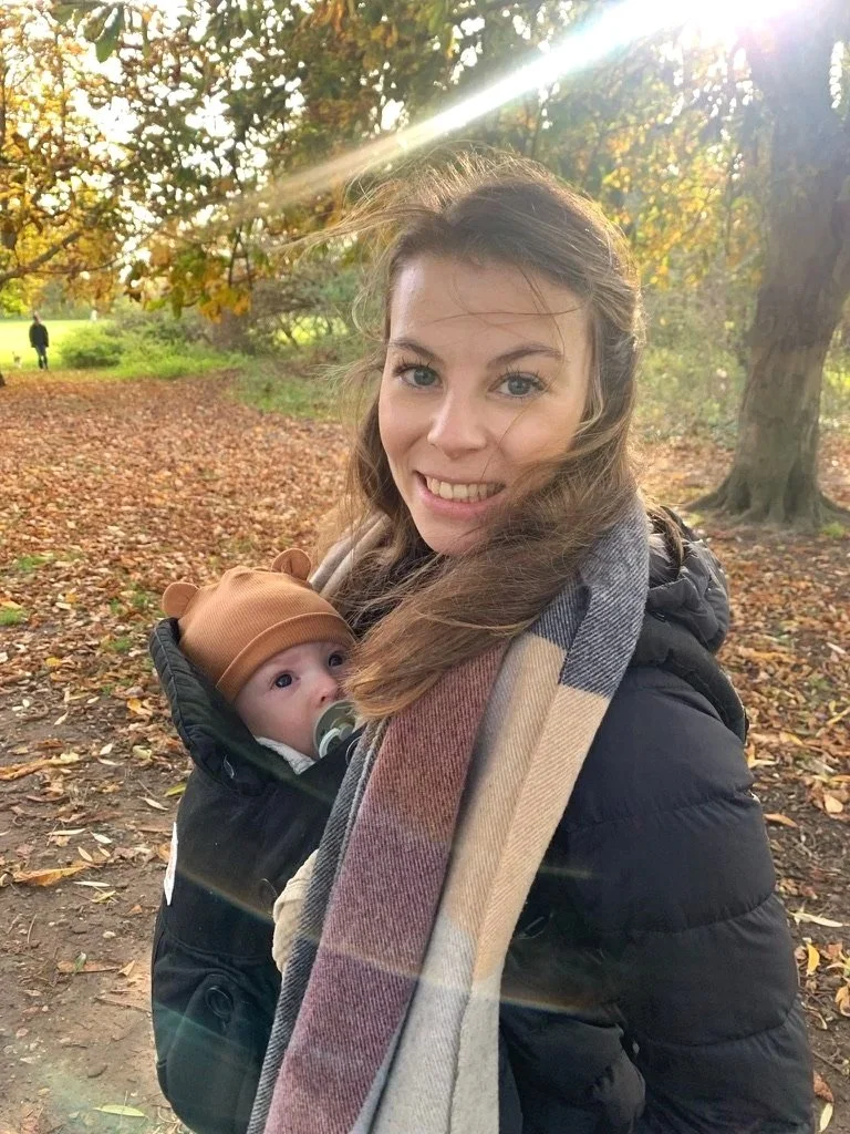 En smilende kvinne bærer en baby i en bæresele på en tur i en skog om høsten, med løv på bakken og fargede trær rundt.