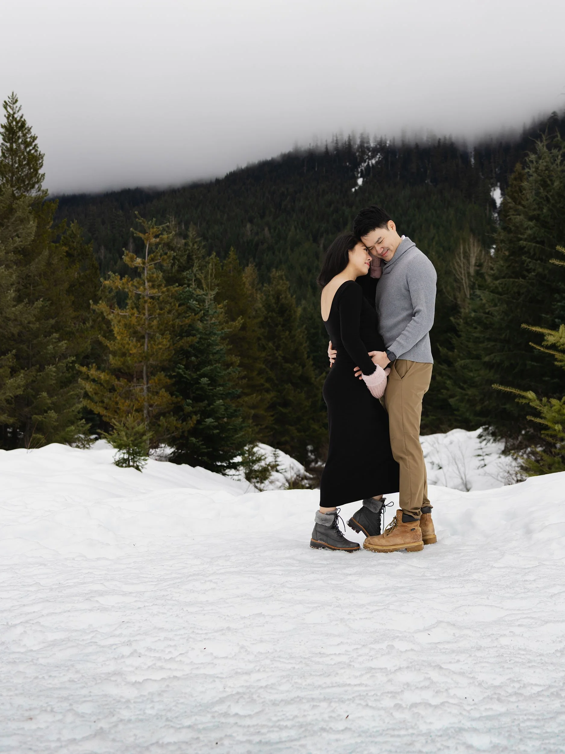 Maternity couple embrace at Gold Creek Pond. Mountains are blanketed in a moody PNW fog. 